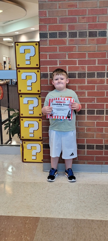 A boy in a green shirt and white shorts holds a certificate in front of a yellow sign with question marks.