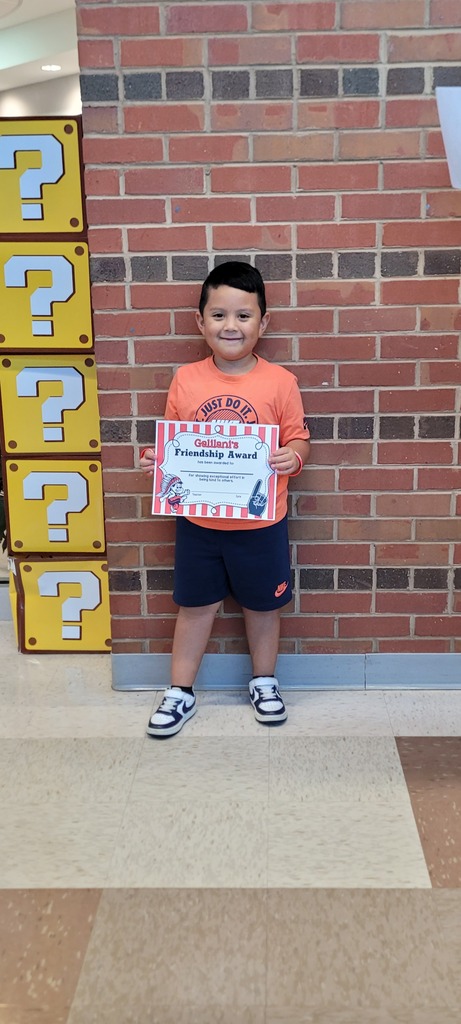 A young boy in orange and blue holds a certificate next to yellow signs with question marks.