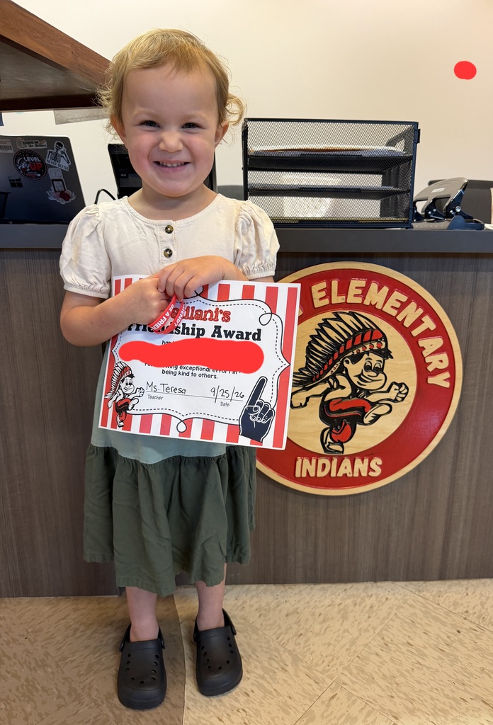 A young child holds a certificate in front of a school sign that reads "Elementary Indians."