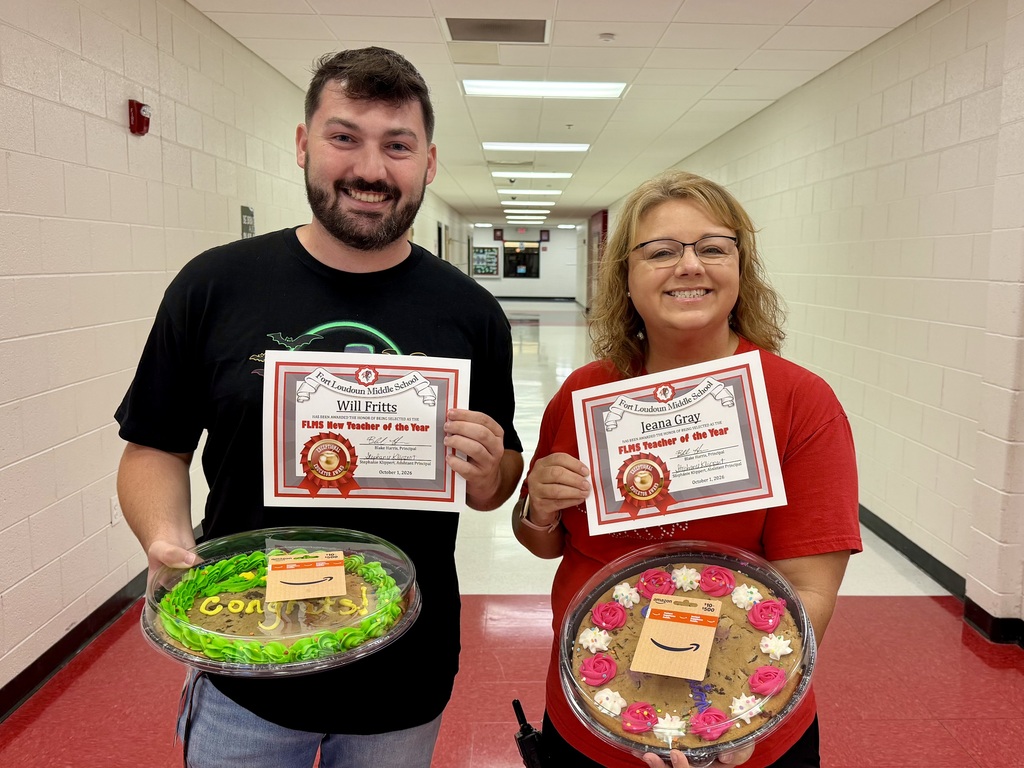 Two people stand in a hallway with a red floor, each holding a certificate and a cake.