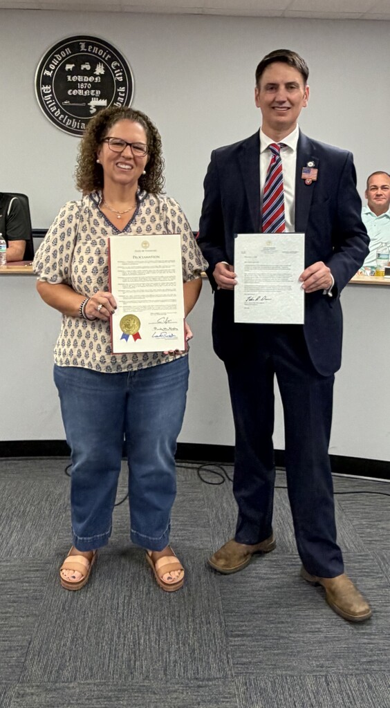 Man and woman stand in a room holding certificates, smiling. They wear formal clothes, and a badge is on the wall.
