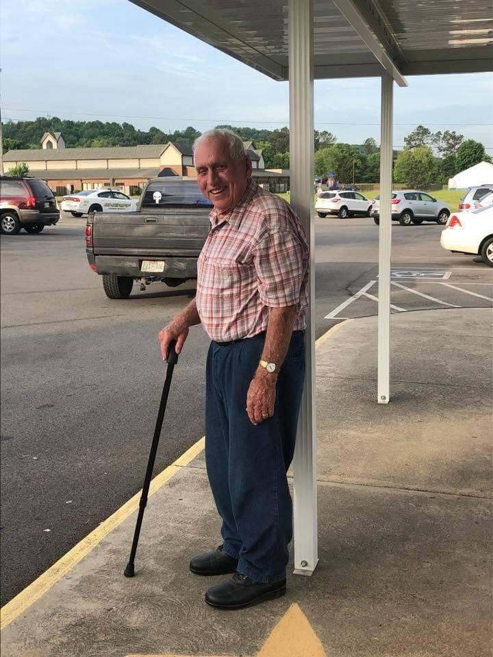 An older man with a cane stands in a parking lot. He wears a plaid shirt, pants, and shoes.