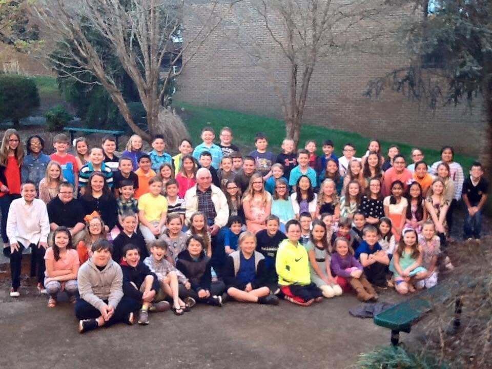 A group of people in varied clothing pose for a photo outside near a building with trees.
