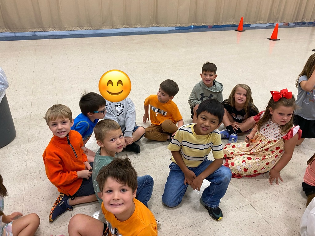 A group of children, some seated and others standing, are in a room. A yellow smiley face emoji is in the center.