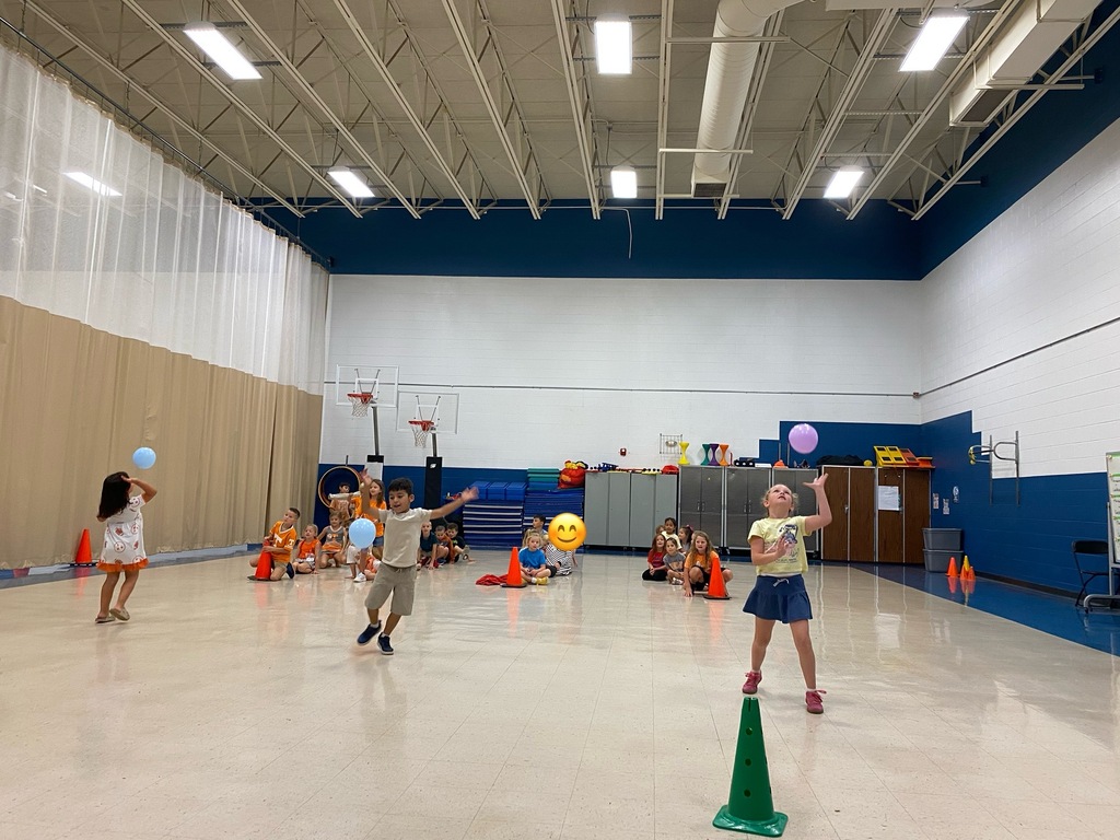 Several children in a gym playing with balloons and cones. The floor is white, and the walls are blue.