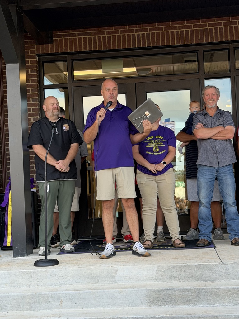 Five adults stand on steps in front of a brick building. One holds a folder and speaks into a microphone.