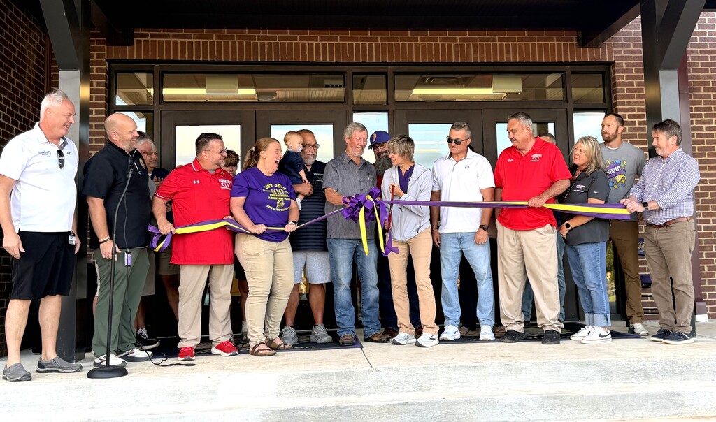 A group of adults, some holding scissors, cutting a purple ribbon in front of a building.