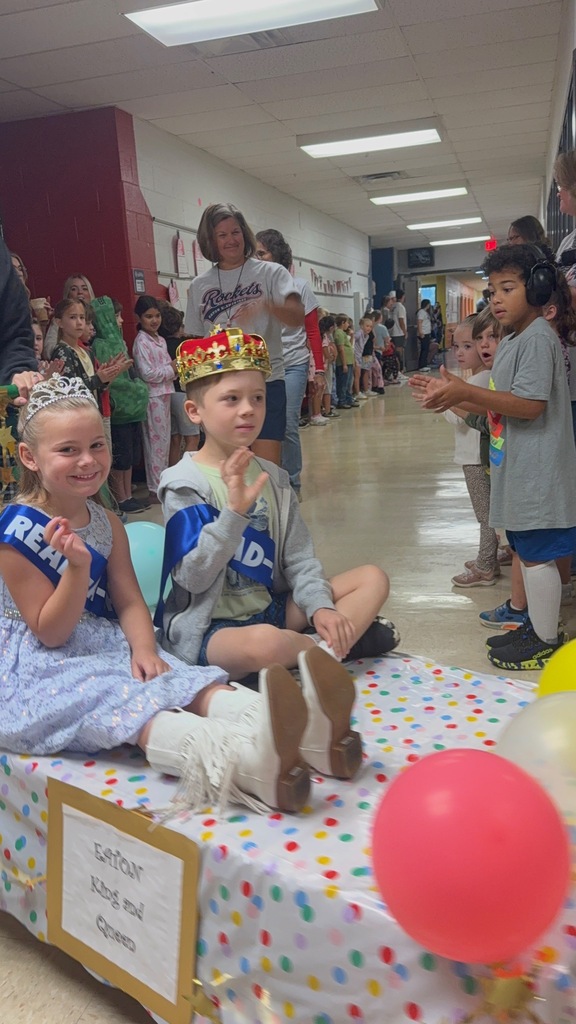 Children seated on a decorated table wearing crowns. One holds a sign that reads "Reign King and Queen."