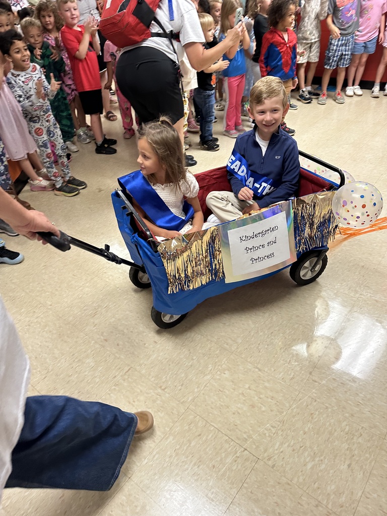 Two children in a decorated cart being pushed by an adult, with others standing in a line.