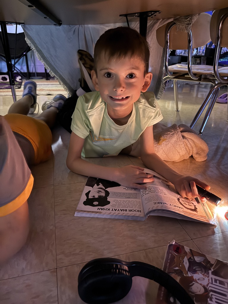 Child lying on floor, smiling, reading book with cartoon character. Headphones and papers nearby.