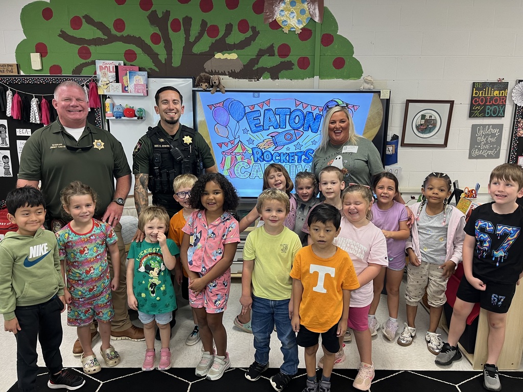 Many children standing with police officers and a woman in a classroom. A large screen displays "Eaton Police" behind them.