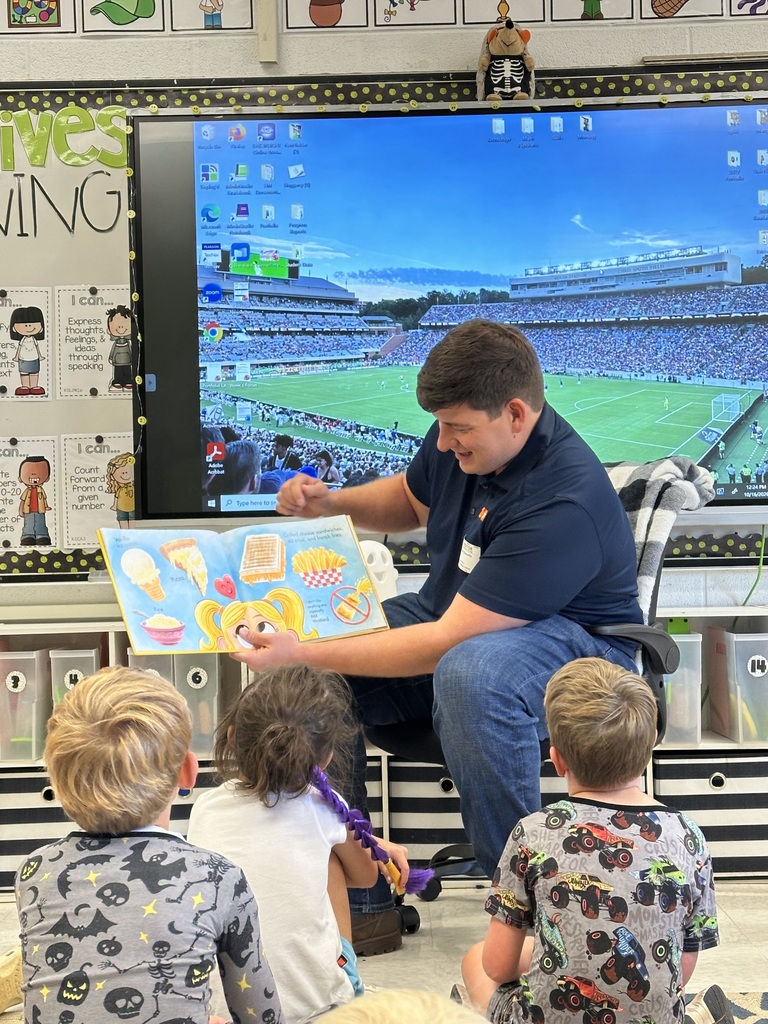 Man sitting in chair reading book to four children. Large screen behind him shows field and seats.