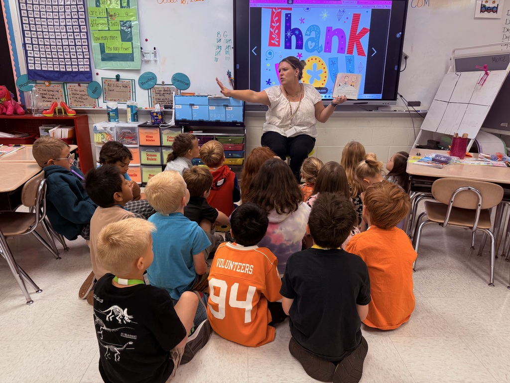 Classroom with children sitting on the floor in front of a woman standing near a screen.
