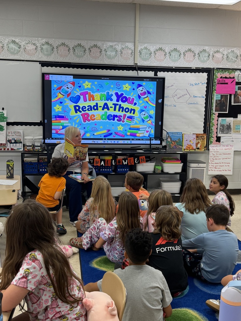 Group of children seated in classroom, watching presentation on large screen. Woman holding book sits on floor.