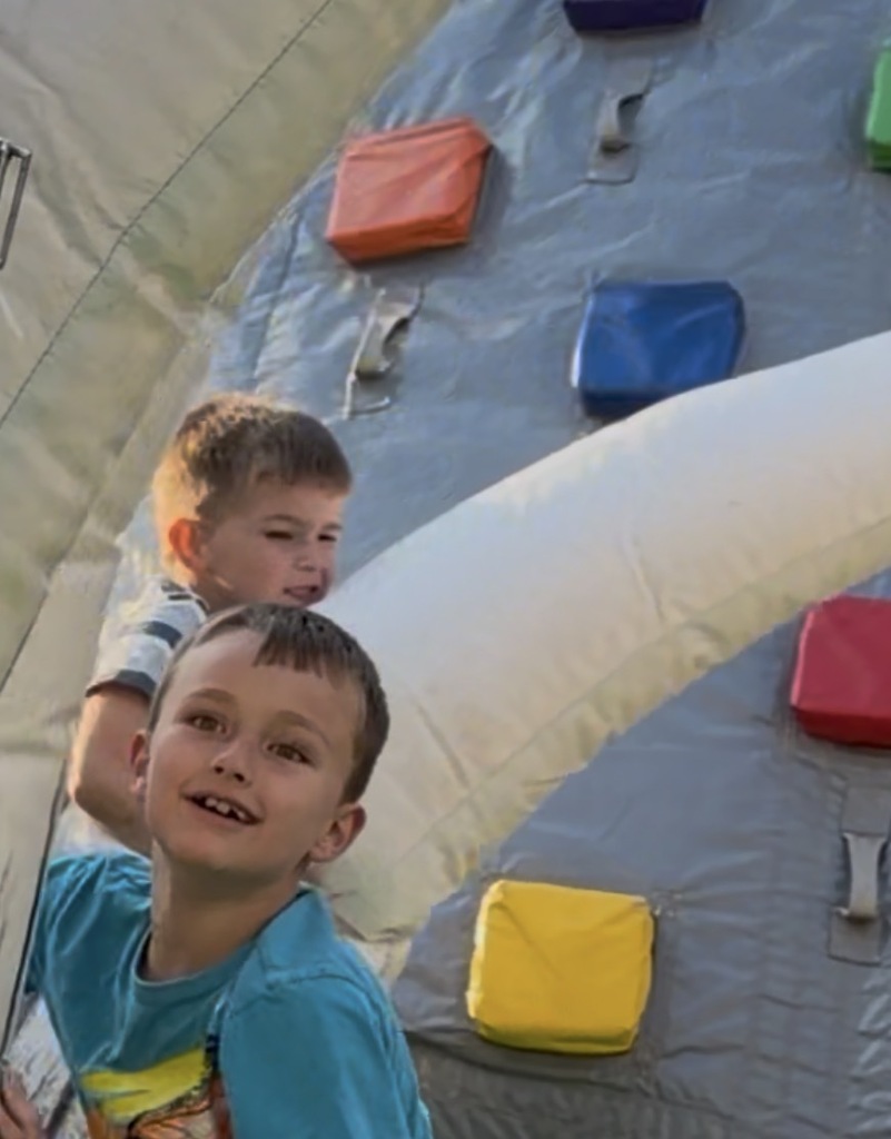 Two boys slide down a blue inflatable slide with colored pouches on either side.