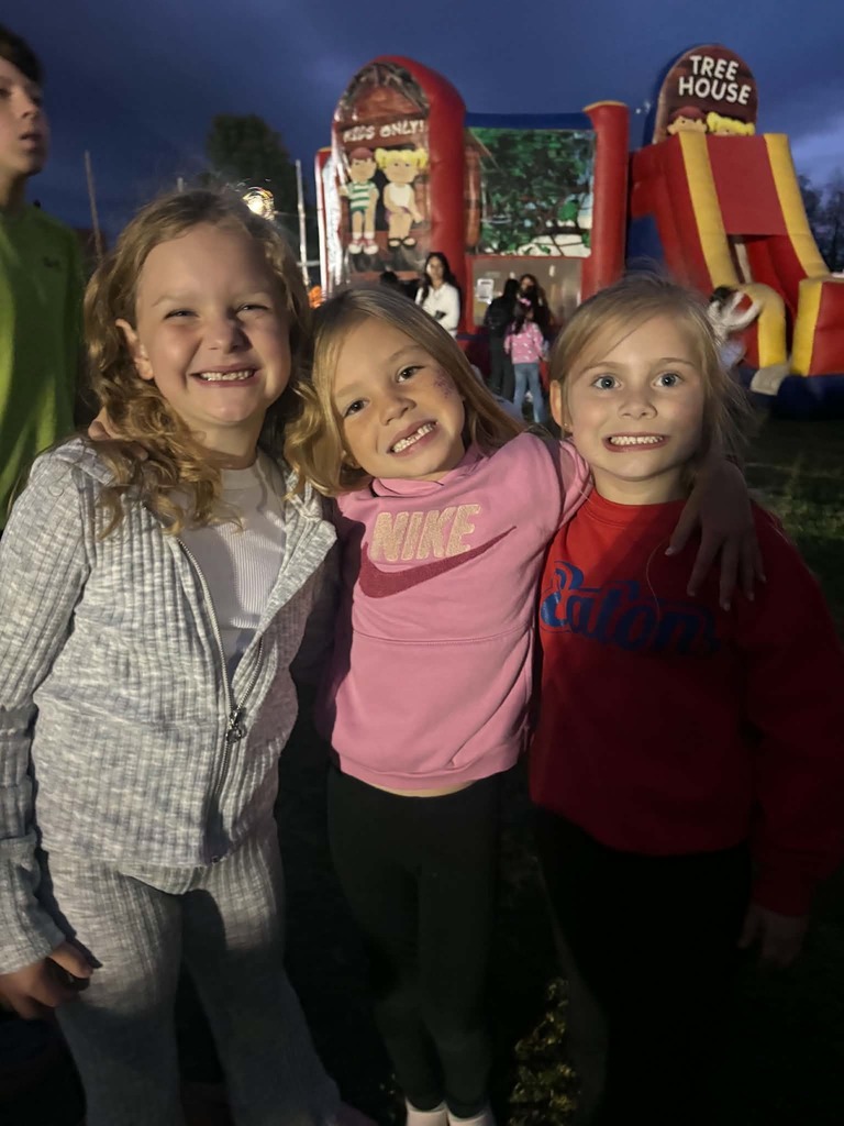 Three young girls stand close together with smiles. Behind them, a person in green stands. In the background, two inflatables are set up.