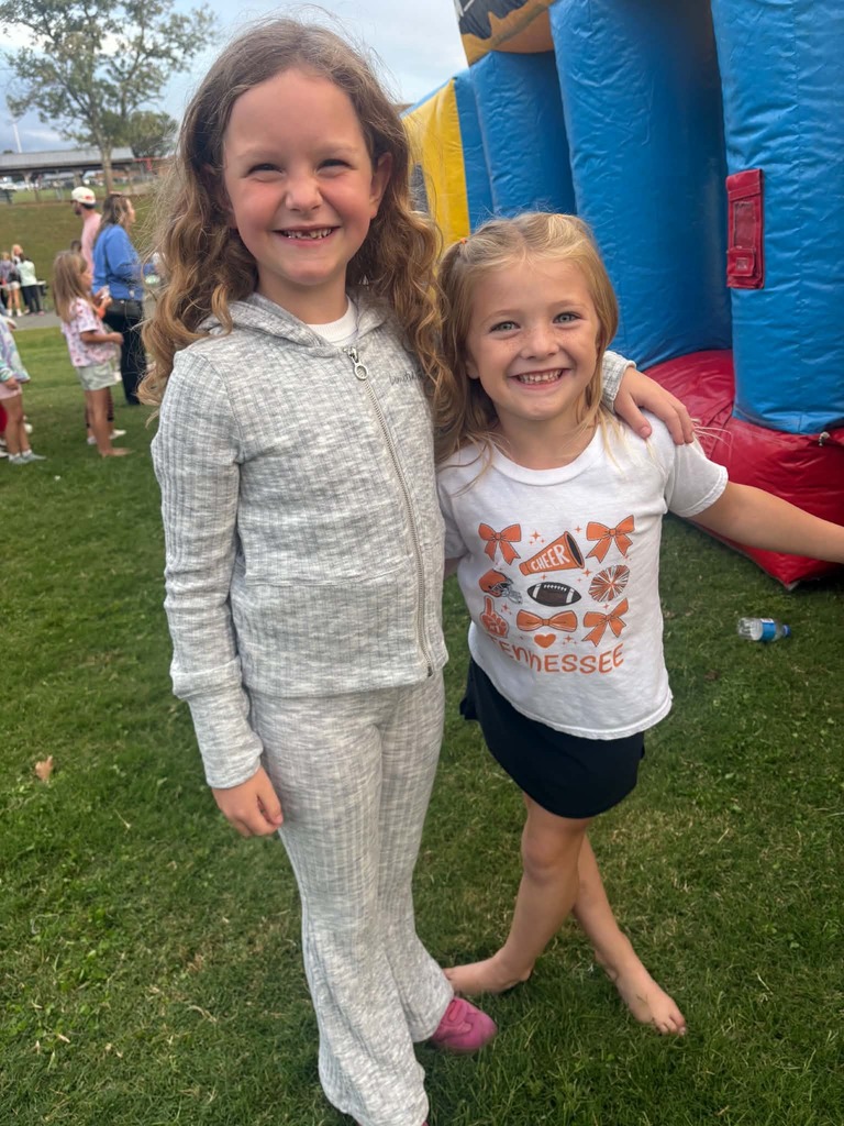 Two girls in matching smiles pose outside near a colorful bouncy house. One girl in a sweatshirt and pants, the other in a t-shirt and shorts.
