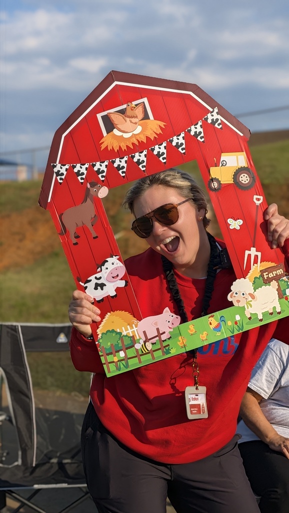 Person in red shirt and sunglasses holds up a red barn cutout with farm animals. Person sits behind with hand on knee.