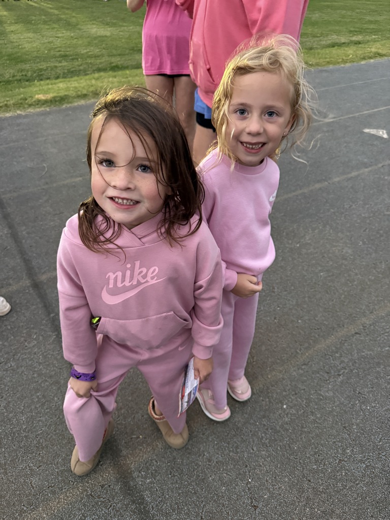 Two young girls in pink Nike outfits smile at the camera on a paved area with grass.