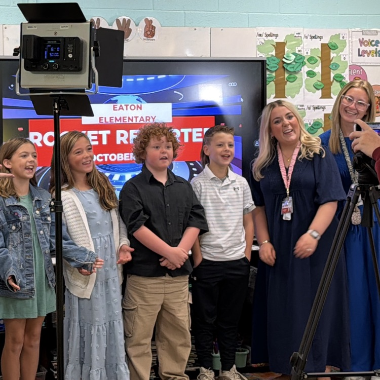 Children and adults standing before a video camera. Monitor shows Eaton Elementary. One woman holds a microphone.