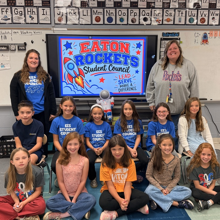 A group of students wearing blue shirts are sitting in a classroom, with three adults standing behind them.