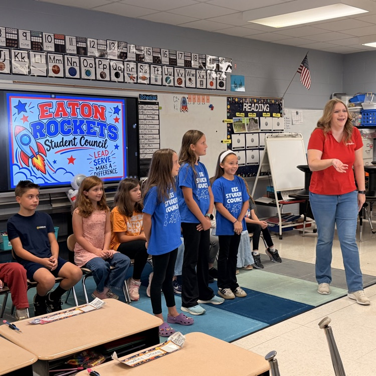 A classroom with a large screen, children wearing blue shirts standing in line, and an adult in red.