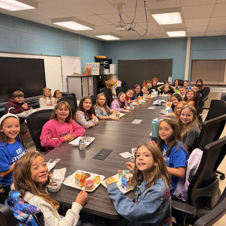 Students in a classroom sit at a long table, eating meals and drinking from cups.