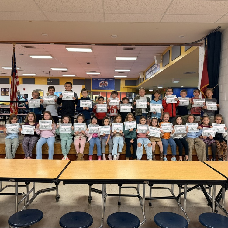 Group of children holding certificates, seated on stage, in front of tables and chairs.