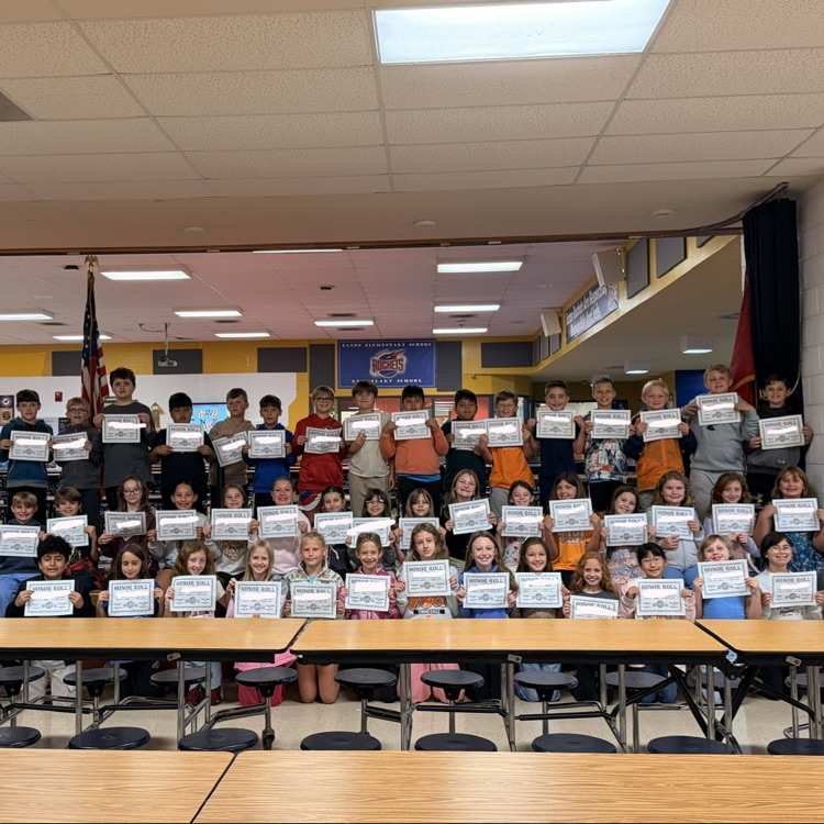 A group of students holding certificates in a classroom with tables, chairs, and flags.