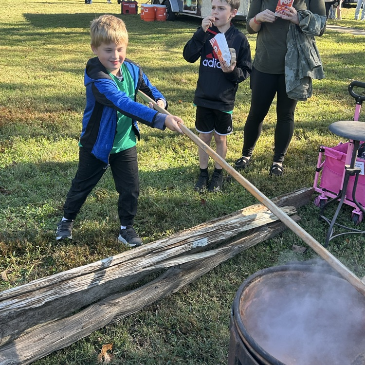 Three children hold wooden sticks, one blows on a fire. One child drinks from a cup.