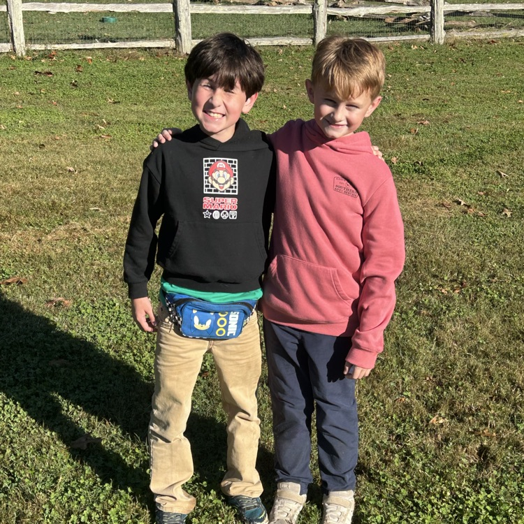 Two boys standing on grass, one wearing a black hoodie, the other a red hoodie. Both wear pants and sneakers. Behind them, a white fence.