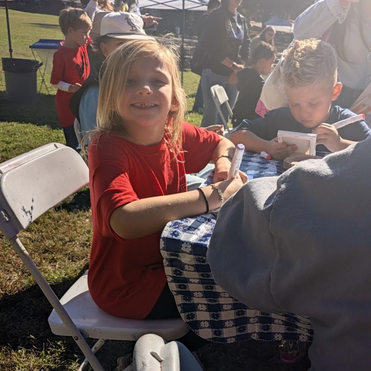 Children seated at tables on grass; girl in red writes with pen, boy in blue reads paper.