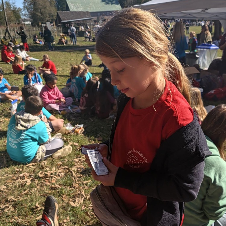 A girl with blonde hair reads a book. Many children sit on grass. A building stands in the distance.