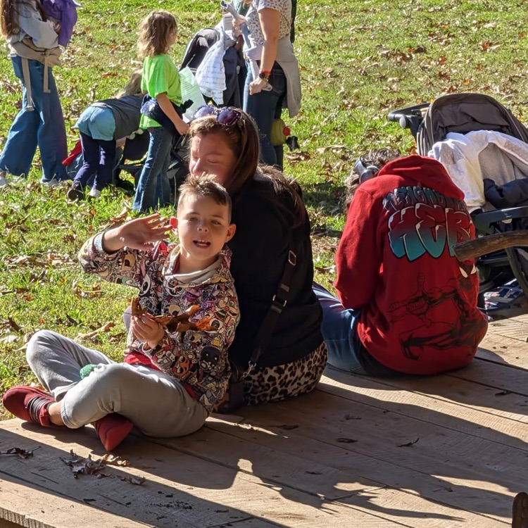 Family with children sitting on wooden bench in outdoor grassy area with strollers. Child wearing floral jacket sits with arms around child in red jacket.