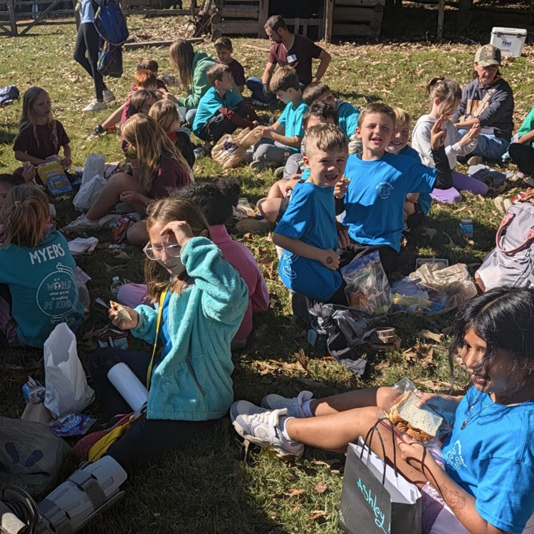 Several children sitting in a circle on grass, some wearing blue shirts and holding food and bags.
