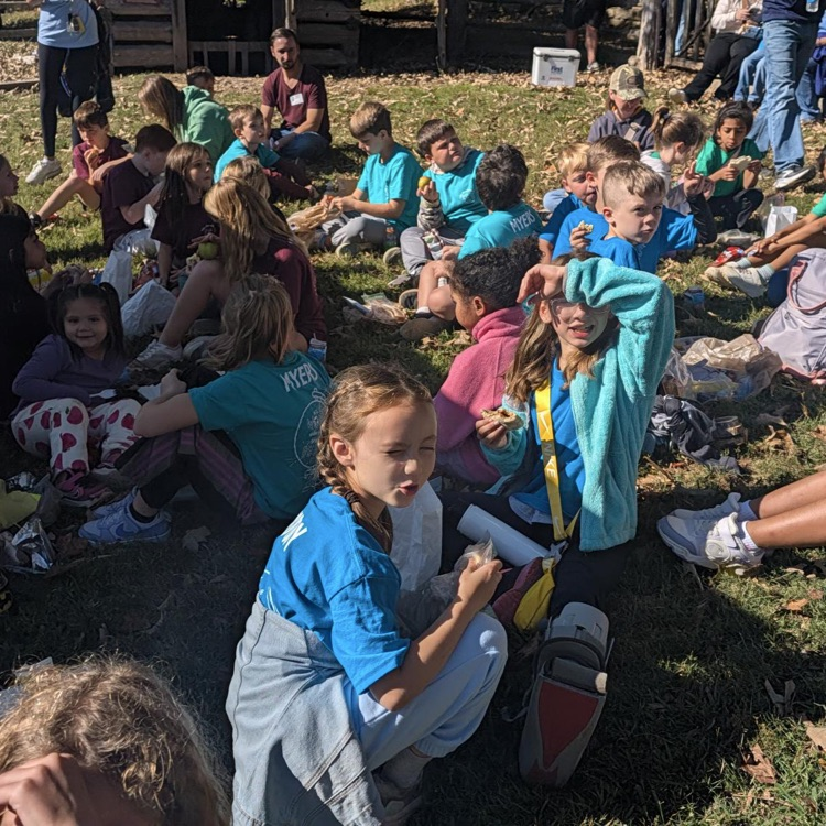 Children sit on grass, some holding food. A girl with glasses and a backpack sits nearby.