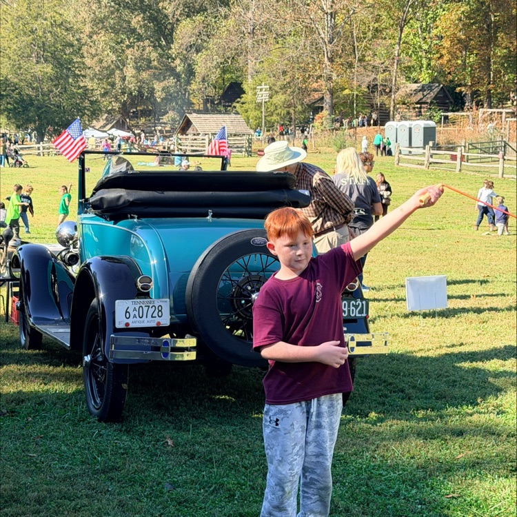 Boy with red hair holding a bird on his arm stands in grass, near an antique car.