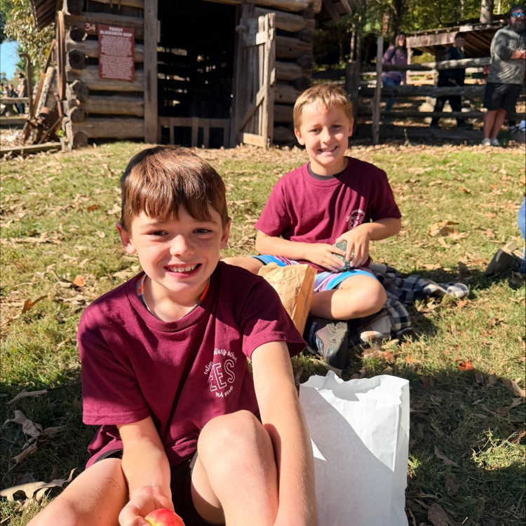 Two boys in maroon shirts sitting on grass near a log cabin, one holding an apple.