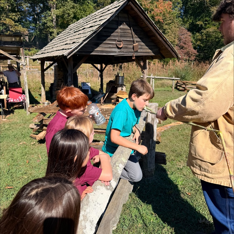 People gather around a small building. A man holds a rope as children lean over a fence.