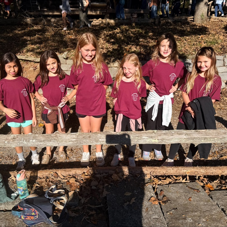 Seven children in matching shirts standing behind a wooden fence with bags and shoes nearby.