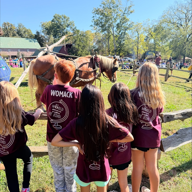 Six students in maroon shirts watch horses standing next to a cart, with a wooden fence and trees in the background.