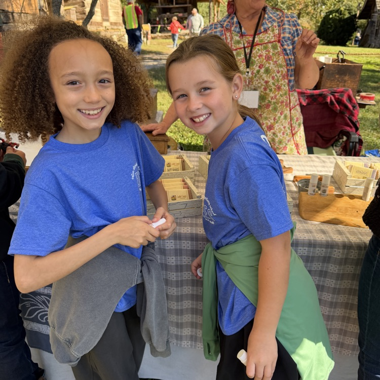 Two girls wearing blue shirts, one with a green apron, stand smiling with a table behind them.