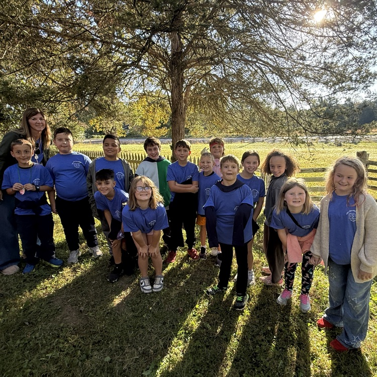 A group of young children in matching blue shirts, standing in a grassy field with trees and a fence in the background.