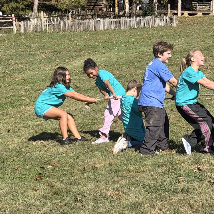 Children in blue and pink shirts holding rope in a grassy field. Trees and a wooden fence in the background.