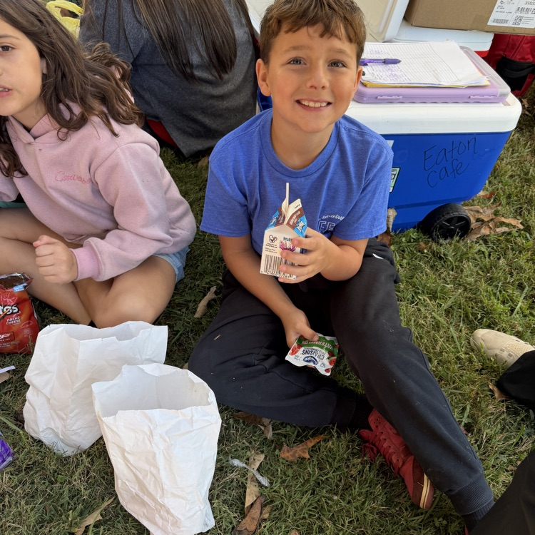 Two children seated on grass, one holding food item, surrounded by bags and storage boxes.