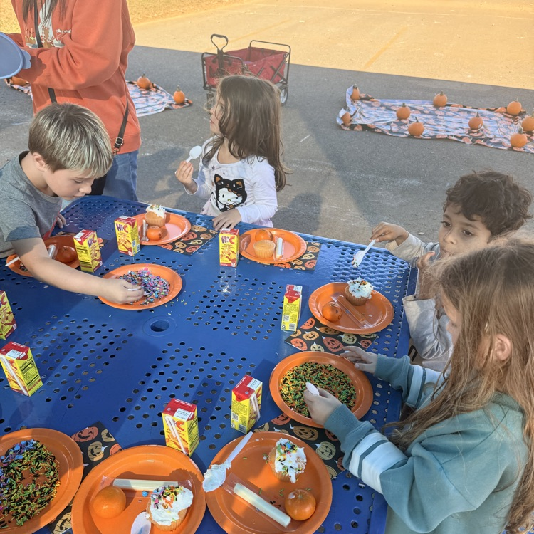 Four children eat at a blue table with orange plates and spoons. Two adults stand behind them.