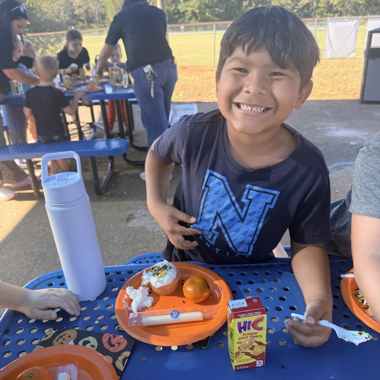 Child smiles at camera, seated at table, holding food items. Blurry adults and children in background.
