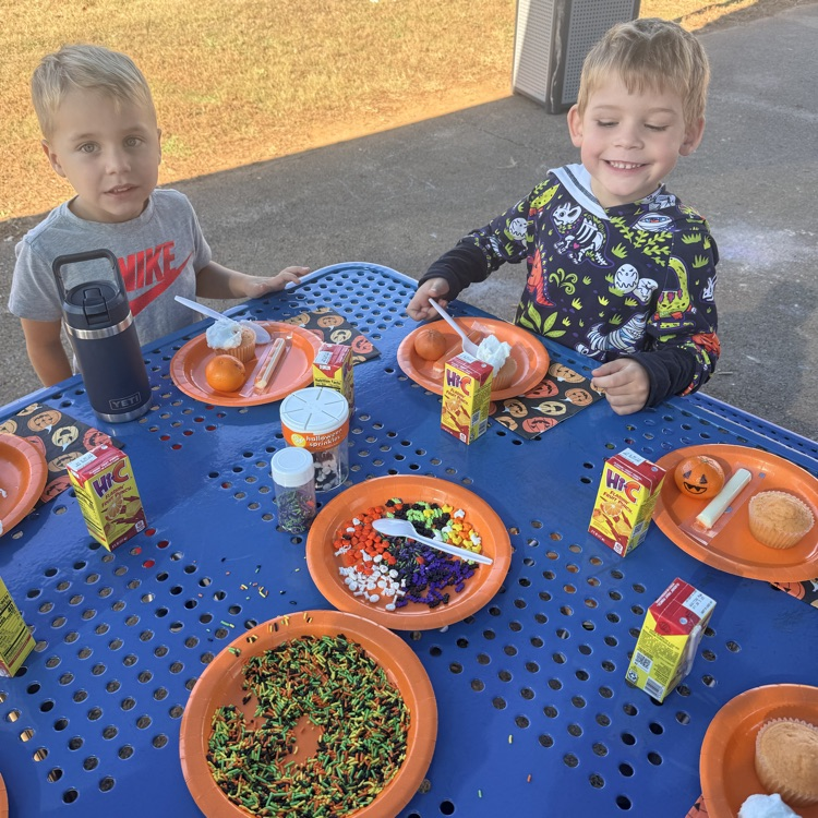 Two children at a table with plates, food, and a container of candy. A sidewalk is behind them.