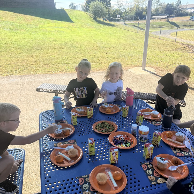 Children seated at a table with food on blue plates, some wearing glasses, in a grassy outdoor area.