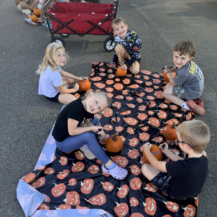 Six children sit around a blanket with pumpkins. They are carving into the pumpkins.